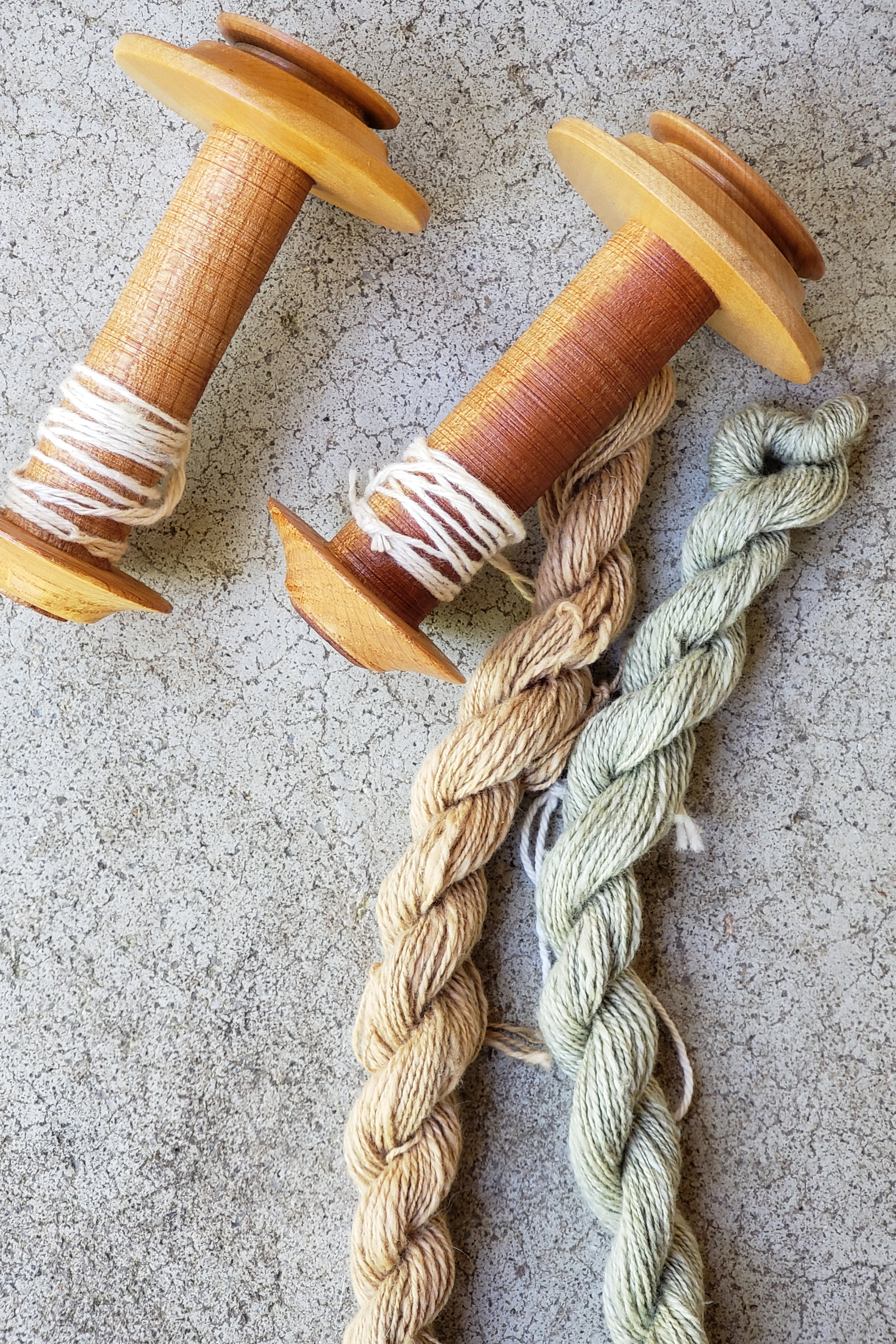 Two mini skeins of handspun naturally colored cotton rest on a concrete background next to two wooden bobbins.